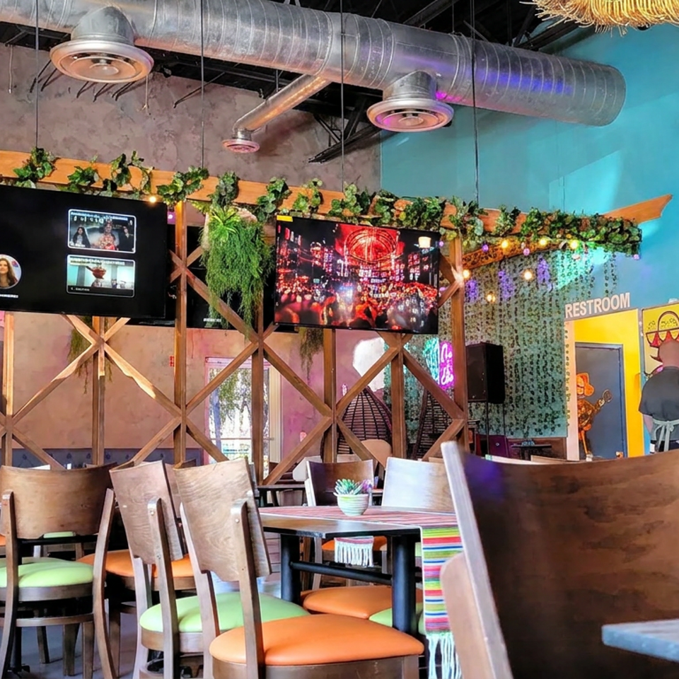 Colorful restaurant dining room with wooden tables, green and orange chairs, hanging plants, televisions, and a restroom entrance.