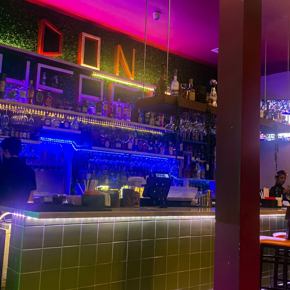 Vibrantly lit restaurant bar with liquor shelves, bartenders serving guests, and diners seated at high-top tables.