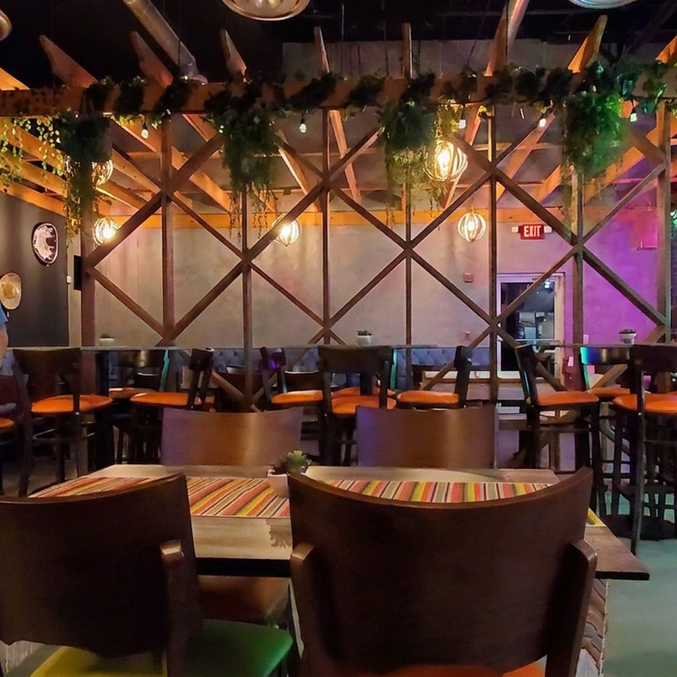 Colorful restaurant interior with patterned seating, hanging greenery, warm pendant lights, and two staff members near the dining area.